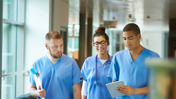 Three healthcare workers in medical scrubs walking together in a hospital hallway while holding documents and a tablet.