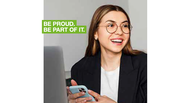 Professional office-style image of a person sitting at a desk, holding a smartphone beside a laptop, with a green text box on the left displaying the message “BE PROUD. BE PART OF IT.”